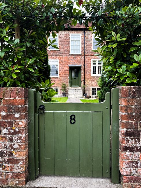 A green wooden garden gate with the number 8, set between two red brick walls with visible weathering and moss, leads into a well-maintained backyard of a traditional brick house. Behind the gate, a narrow stone pathway bordered by a lush, neatly trimmed lawn extends towards the front door, which is dark green and framed by white window frames with multiple panes. Flanking the pathway are garden beds with a variety of leafy plants and shrubs, some with glossy dark green leaves, contributing to a tidy and inviting outdoor space. Mature trees and hedge borders create a sense of privacy and enclosure, while the overall environment appears lush and cared for, typical of urban gardens on a council estate in the E8 postcode area. The scene is captured in natural daylight, highlighting the textures of the bark, brickwork, and foliage, emphasizing the well-maintained outdoor area that Gardeners Hackney would be pleased to support with garden maintenance and landscaping work in such settings.