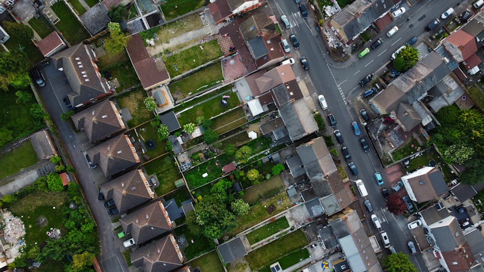 An aerial view of a residential area on a council estate with terraced houses and small gardens. The gardens are divided by fences and feature textured lawns, flower beds, and patches of shrubbery. Some gardens have patio areas with outdoor furniture, while others contain vegetable patches or decorative elements. The streets are lined with parked cars, and the roads are bordered by grassy verges and sidewalks. Several trees and hedges provide greenery to the scene, and the lighting appears to be natural daylight, highlighting the different textures of grass, soil, and foliage. Visible gardening tools like rakes or shears are not present, but the overall scene suggests regular garden maintenance and landscaping work that Gardeners Hackney can assist with in E8 postcode council estate gardens.