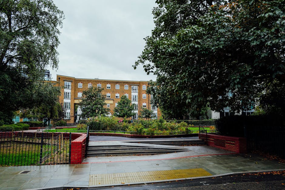 The image depicts an urban garden situated in front of a multi-story brick residential building near Hackney Central station. The garden area is enclosed by a black metal fence with a red brick base, featuring a small set of concrete steps leading up to a paved pathway. Surrounding the pathway, there are well-maintained flower beds with a variety of green plants, shrubs, and small flowering plants. Mature trees with lush green foliage provide partial shade over parts of the garden, creating a serene environment amidst the cityscape. The ground in the garden is a mix of grass and soil, appearing moist and freshly tended, indicating recent garden maintenance or landscaping work. The scene is captured under overcast skies, giving a soft, diffused light that highlights the textures of the trimmed grass, leaf foliage, and the neat edges of the garden beds. This setting exemplifies routine lawn care and hedge trimming practices carried out by Gardeners Hackney, who specialize in garden maintenance for homes near Hackney Central station.