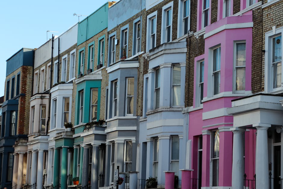 A row of Victorian terraced houses in Stoke Newington, each featuring a bay window on the ground floor and tall, narrow sash windows on the upper floors. The houses are painted in pastel shades of blue, green, white, and pink, with some displaying decorative details around the windows and on the cornices. The facades show a mix of brickwork and smooth painted surfaces, with small front gardens enclosed by low black metal fences. The gardens contain plants, shrubs, and potted flowers. The sky above is bright and clear, casting soft natural light that highlights the textures of the brickwork, painted surfaces, and plants, reflecting a typical residential street scene. The overall scene captures the charm of Victorian architecture with a focus on well-maintained exteriors, indicative of professional garden and outdoor space maintenance, with Gardeners Hackney providing expert gardening and landscaping work for such properties.